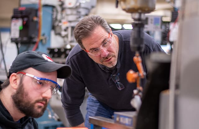 An AJAC instructor works with an apprentice on a manual milling machine.